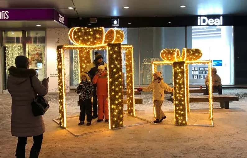 AVM girişinde altın ışıklı hediye kutusu kemerinden geçen aile ve çocuk