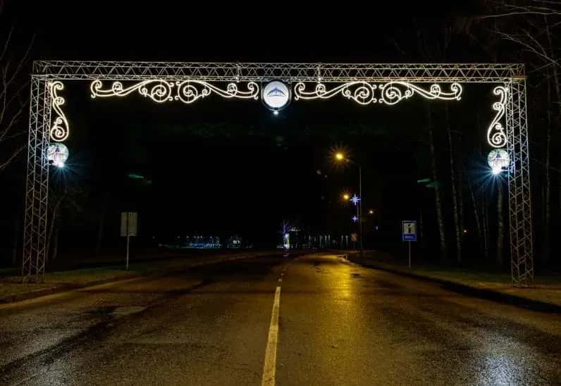 Ağaçlı bir yol girişinde LED tak kemeri, kemer ayaklarında dairesel madalyonlu sarmal direk süsleri