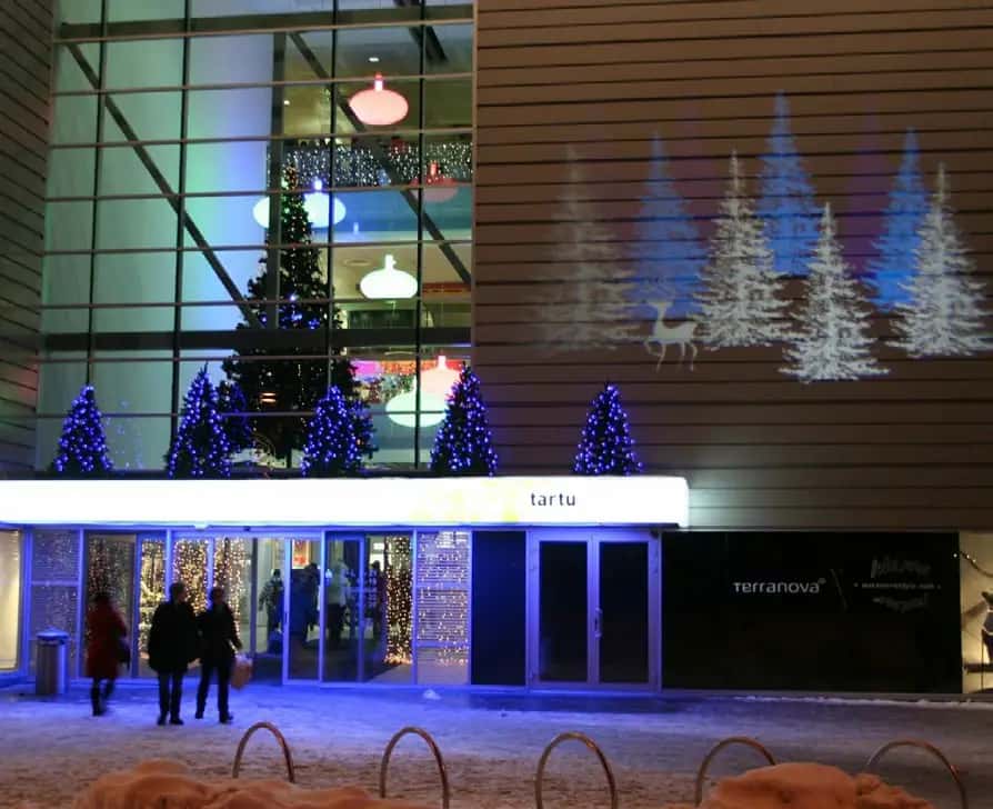 Gece alışveriş merkezi girişinde duvara yansıtılmış beyaz ağaç ve geyik desenleri, mavi ışıklı küçük ağaçlar