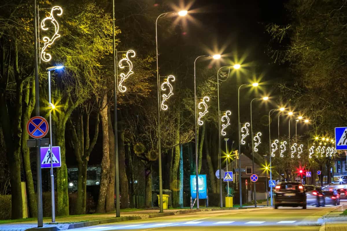 Ağaçlı yol kenarında sıralı direklerde beyaz kıvrımlı motifler, gece