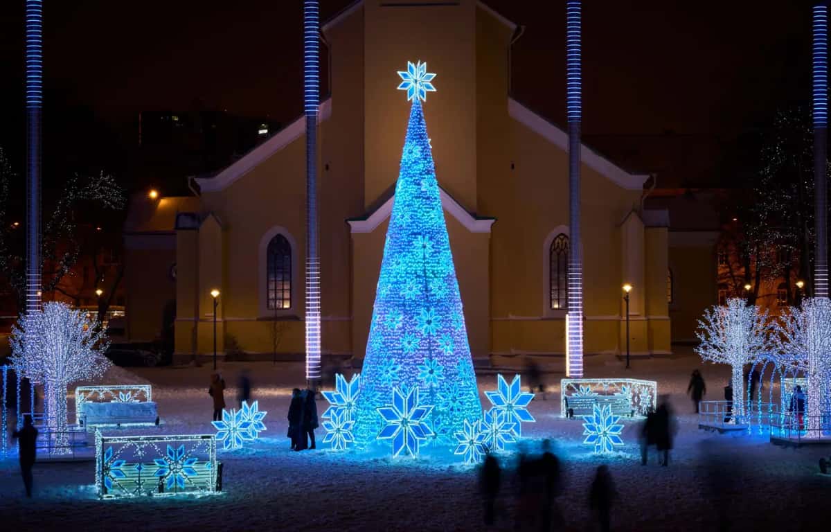 Kilise önündeki meydanda mavi ışıklı koni ağaç, çevresinde kar tanesi figürleri, gece