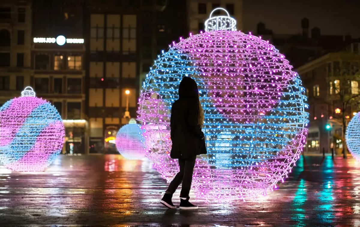 Islak zeminli meydanda pembe-mavi dev LED toplar, önünde yürüyen kadın, gece