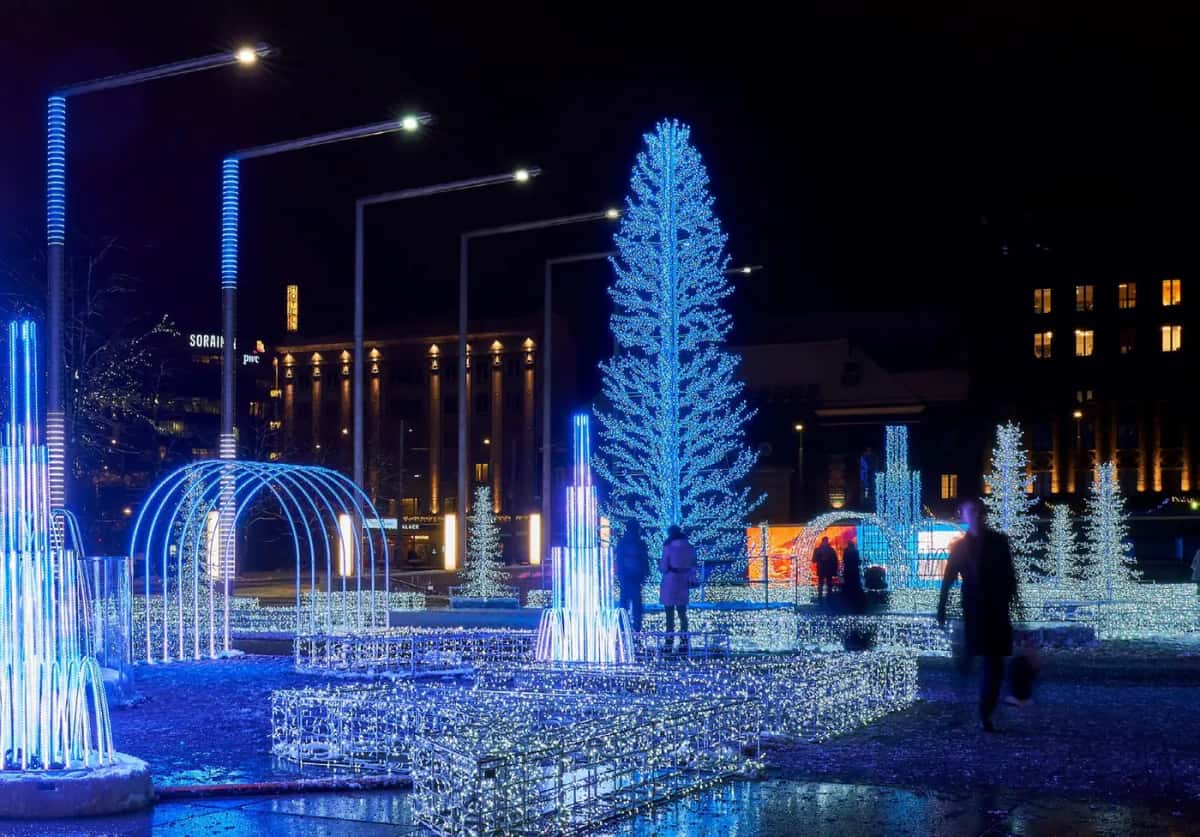 Meydanda mavi ışıklı büyük ağaç, kemerler ve ışık çeşmeleri, arkada binalar, gece