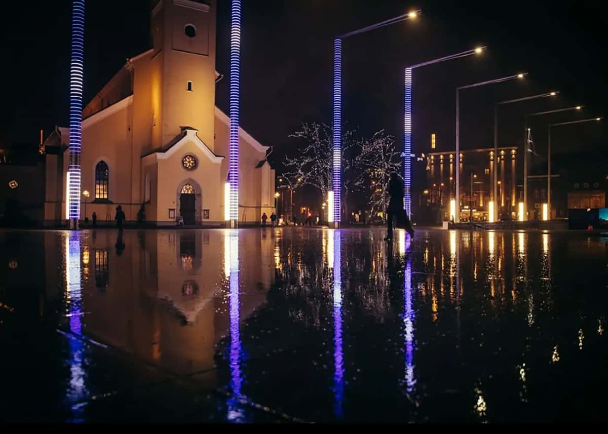 Kilise önündeki ıslak meydanda mor ve mavi LED direkler, yansıması zeminde, gece