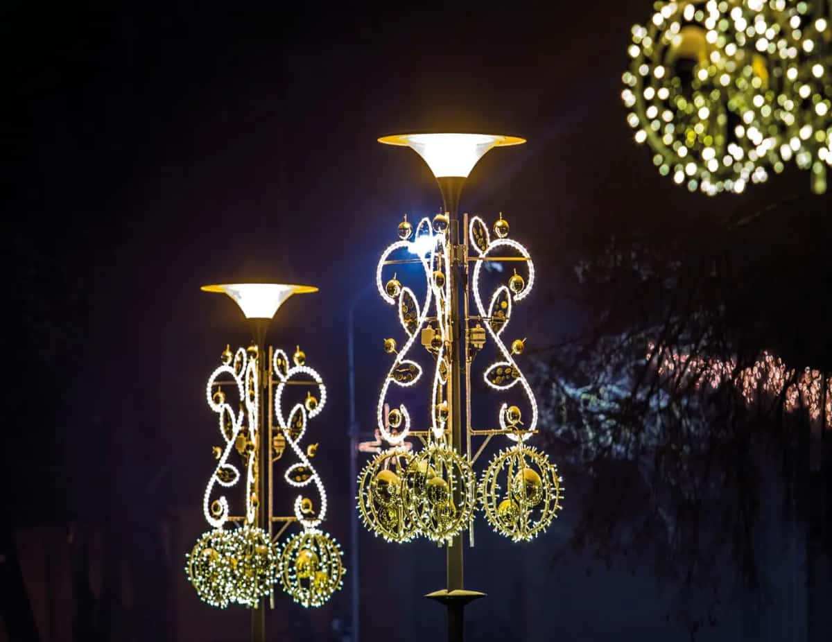 Süslemeli lamba direklerinde kıvrımlı altın LED motifler ve yeşil çelenk toplar, gece