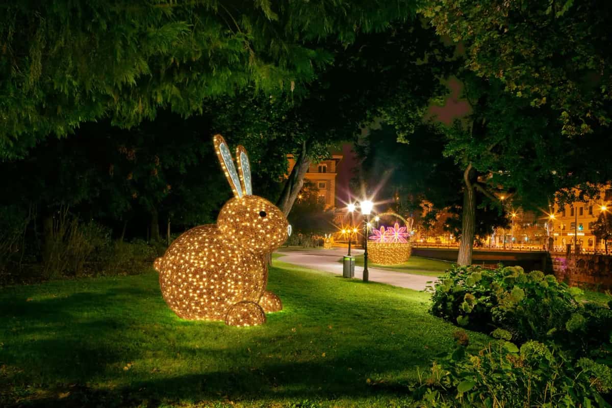 Park çimeninde ağaç altında duran, gümüş kulaklı altın sarısı dev LED tavşan