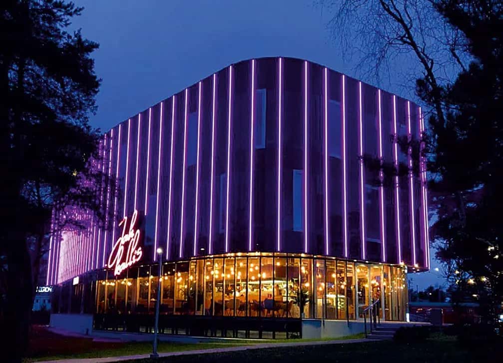 Dikey mor LED çizgilerle aydınlatılmış kavisli cephe, altında camlı restoran katı, akşam